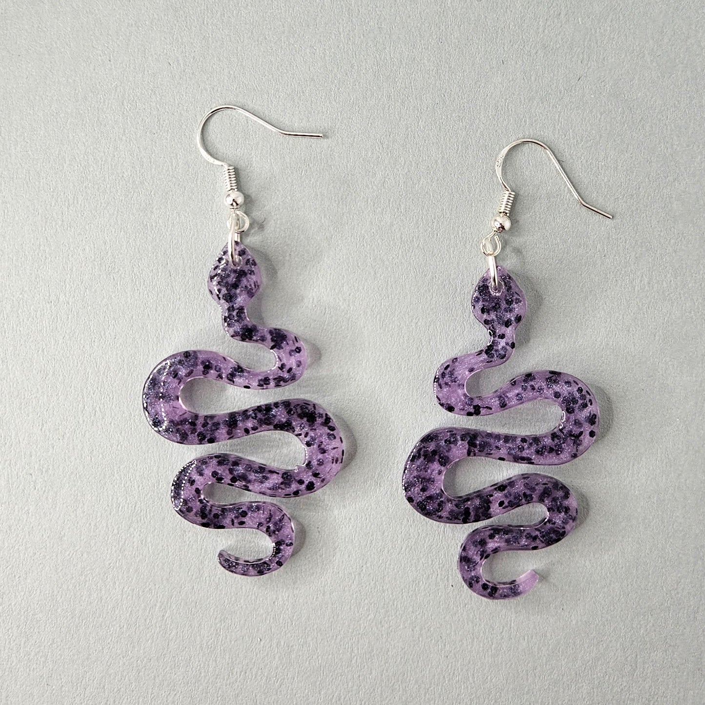 Ein Paar violette Schlangen-Ohrringe aus Resin mit schwarzen Sprenkeln. Die Ohrringe haben versilberte Hakenverschlüsse.
