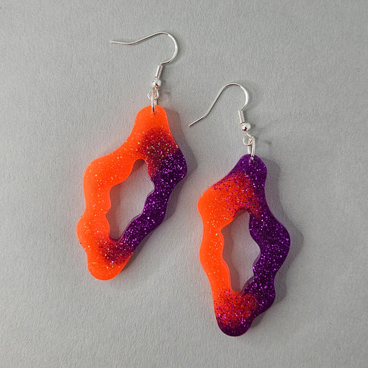 Orange und violette Glitzer-Ohrringe aus Resin mit unregelmäßiger Form und versilberten Hakenverschlüssen.