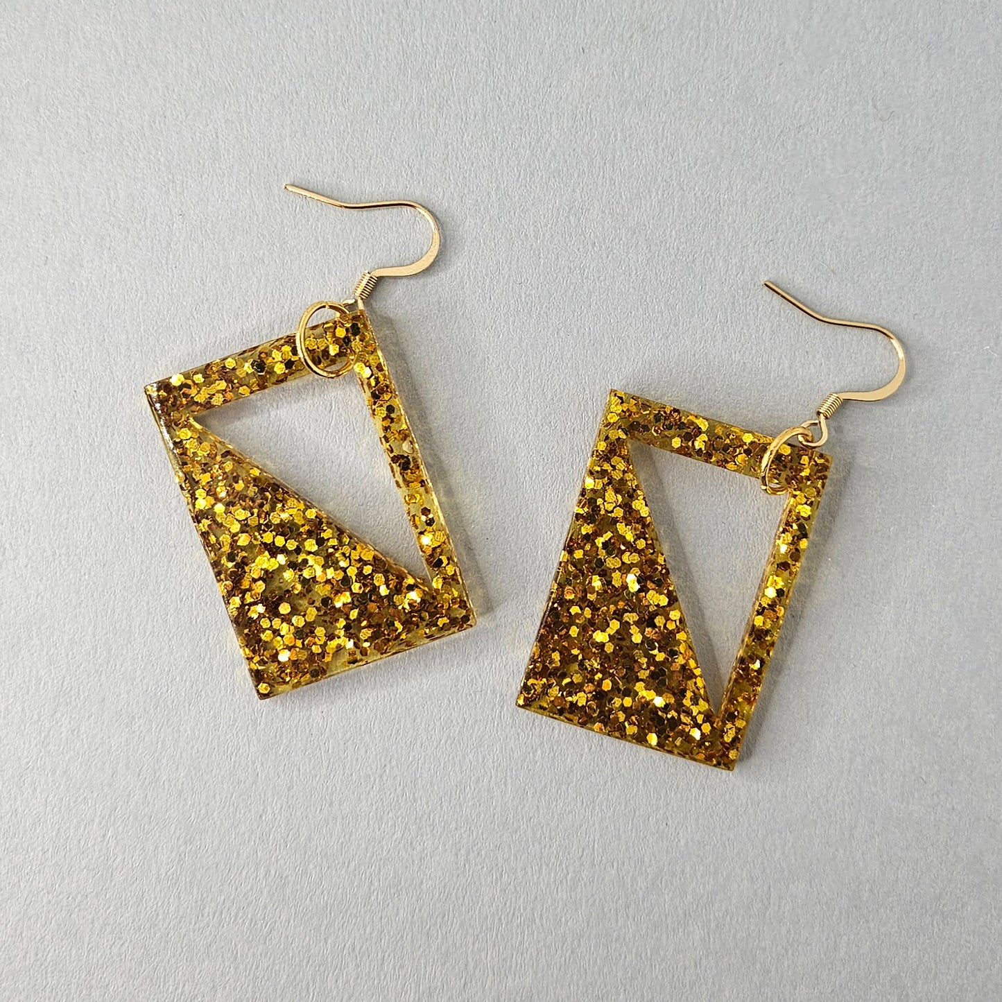 Goldene Glitzer-Ohrringe aus Resin mit geometrischem Design. Die Ohrringe sind quadratisch mit einem dreieckigen Ausschnitt und funkeln durch eingearbeitete Glitzerpartikel. Mit vergoldeten Haken